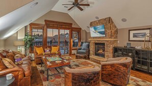 A cozy living room with a fireplace, leather sofas, a coffee table with board games, and a view of a snowy landscape through large windows.