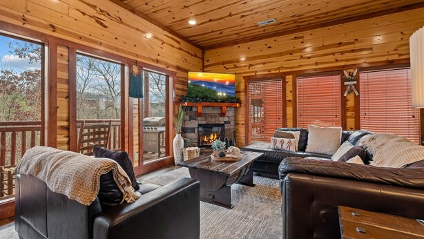 A cozy log cabin interior with a stone fireplace, leather furniture, and large windows offering a view of the outdoors. - Pvt Indoor Pool, Gorgeous 6-BR Cabin, Theater, BBQ (Sevierville)