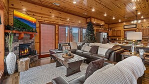 A cozy cabin interior with a fireplace, leather sofas, a dining area, and a Christmas tree. - Pvt Indoor Pool, Gorgeous 6-BR Cabin, Theater, BBQ (Sevierville)