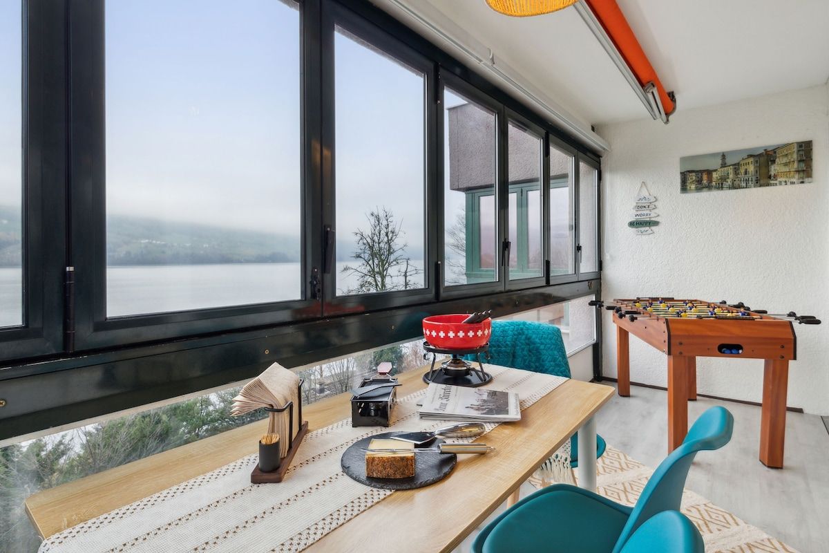 A room with a large window overlooking a body of water, a wooden table with a book and a camera, and a foosball table.