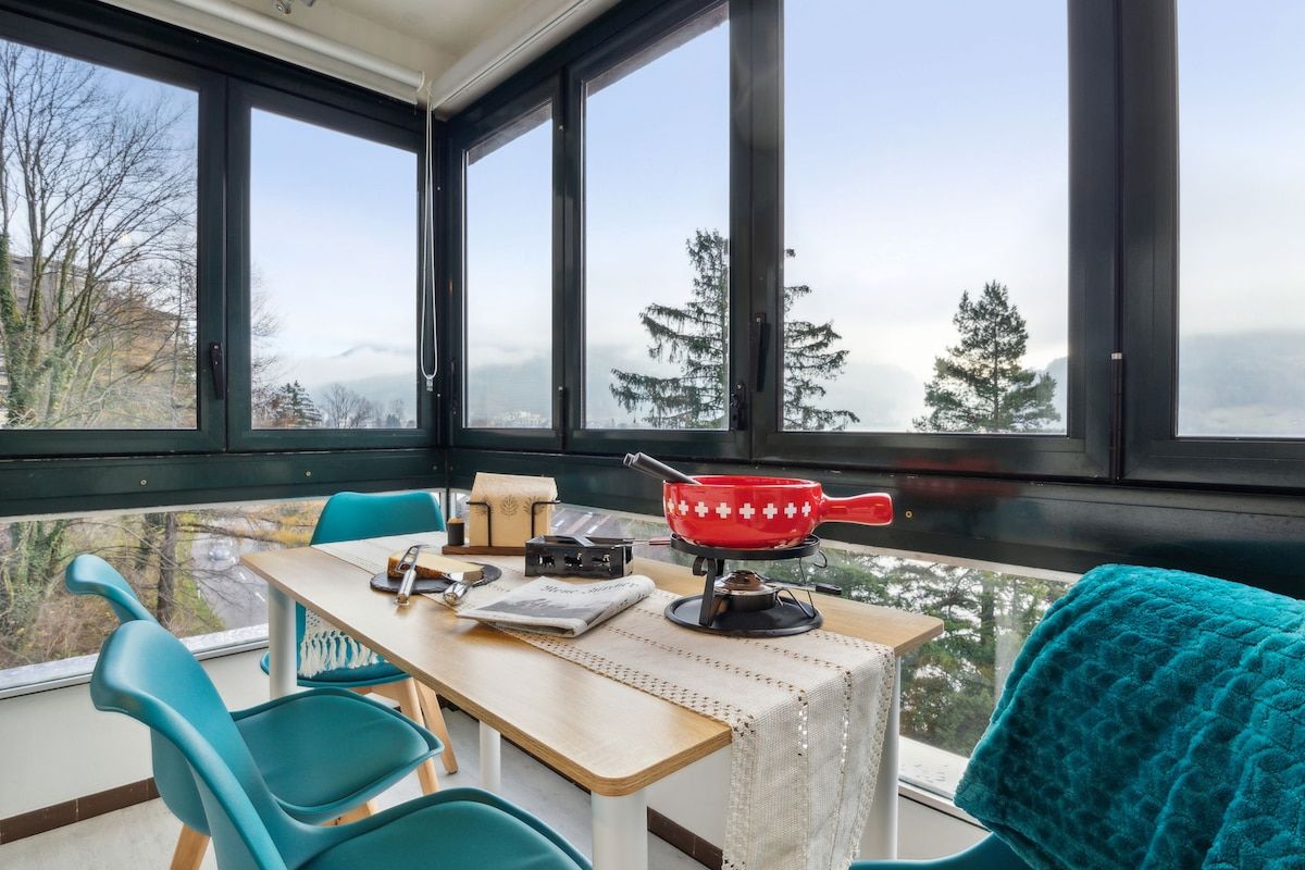 Een kamer met een eettafel, stoelen, een rode pan op een fornuis en uitzicht door grote ramen.