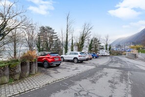 A parking area with several cars and a camper van, surrounded by trees and a mountainous backdrop.