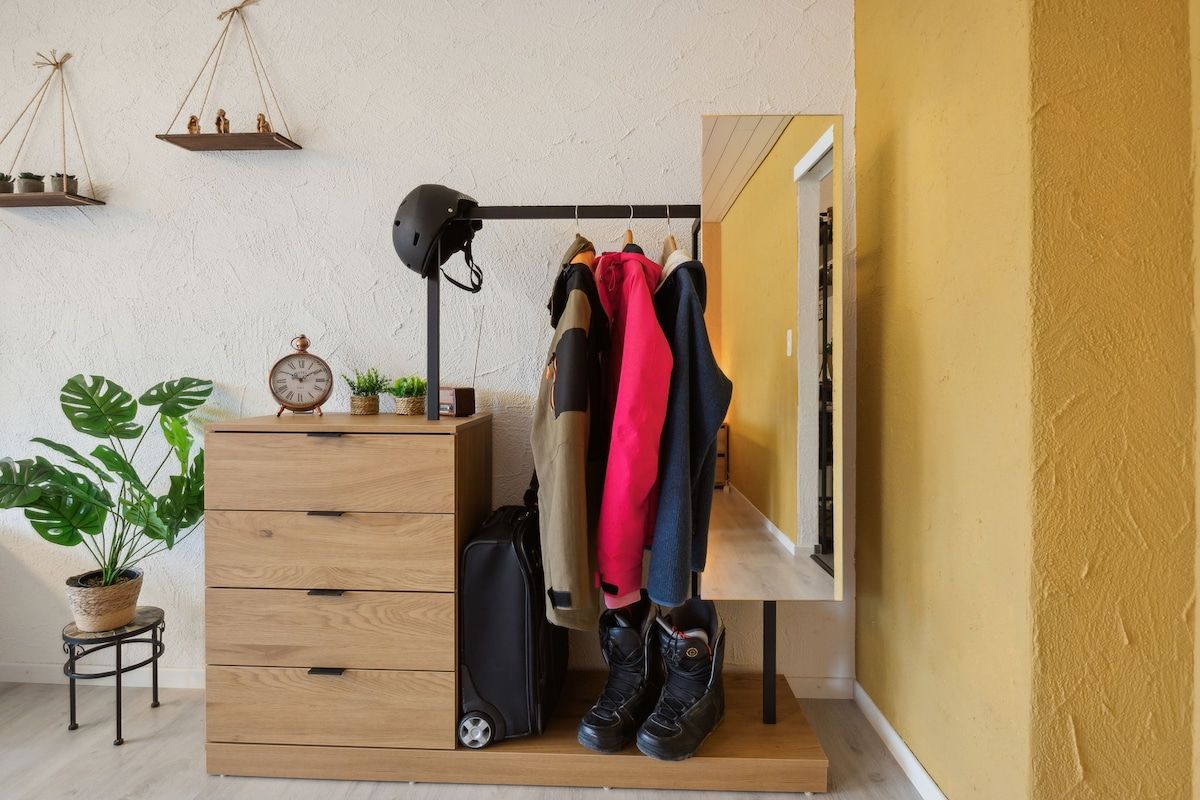A room with a wooden dresser, a hanging coat rack with several jackets, a potted plant, and a suitcase.