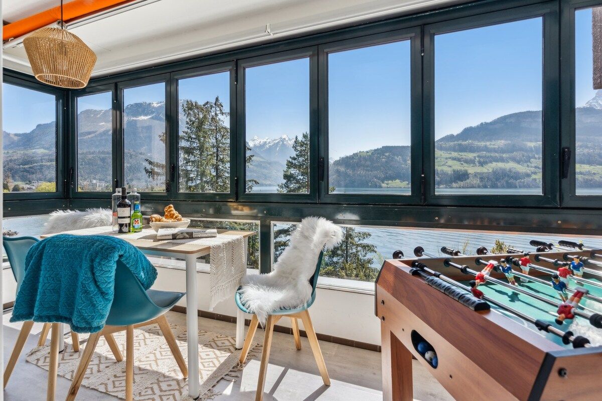 Ett rum med ett fotbollsspel, ett bord med flaskor och en skål med frukt, och utsikt över berg genom stora fönster.