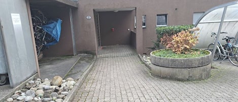 Ein gepflasterter Weg führt zum Eingang eines Gebäudes, ein Betonpflanzgefäß mit Pflanzen und ein Fahrrad, das davor abgestellt ist.