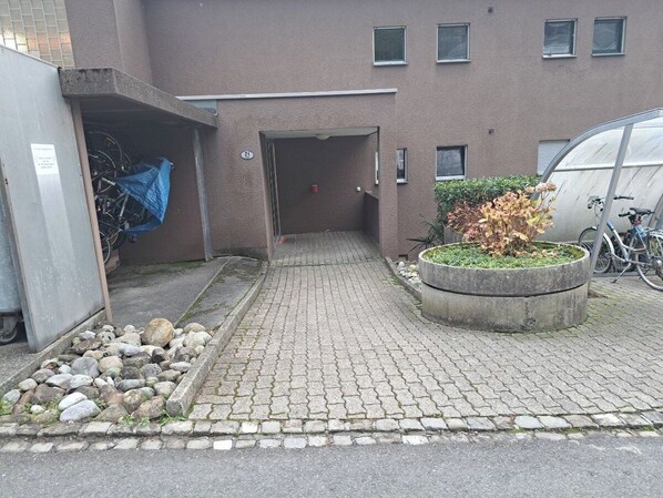 A paved pathway leading to a building entrance, a concrete planter with plants, and a bicycle parked outside.