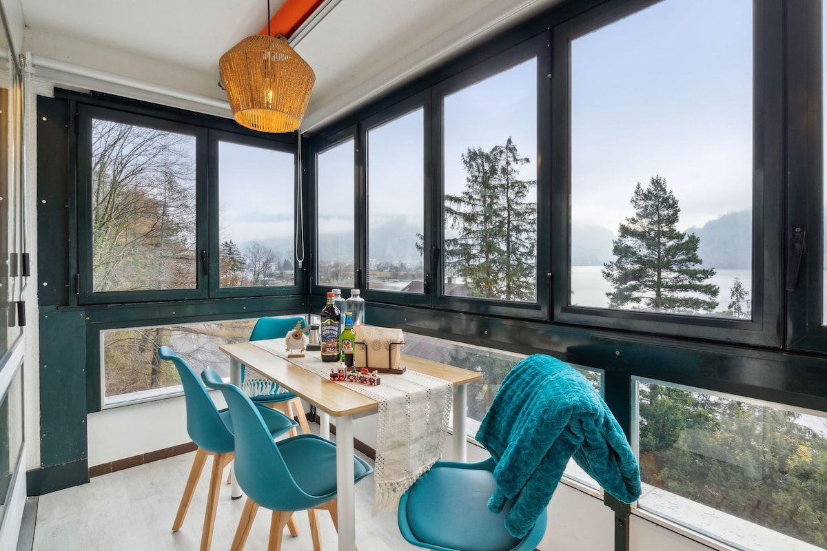 Ein Zimmer mit großem Fenster, das auf einen Wald und einen See blickt, ein Tisch mit Stühlen und einer Hängelampe.