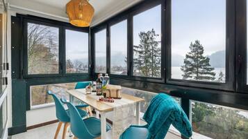 Ein Zimmer mit großem Fenster, das auf einen Wald und einen See blickt, ein Tisch mit Stühlen und einer Hängelampe.