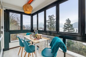 A room with a large window overlooking a forest and a lake, a table with chairs, and a hanging light fixture.