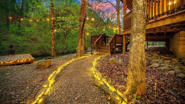 Una cabaña de madera con terraza, rodeada de árboles y un camino de grava con luces colgantes.