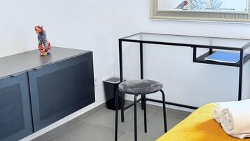 A modern room with a grey sofa, a small stool, a desk with a chair, and a bed with a yellow blanket.
