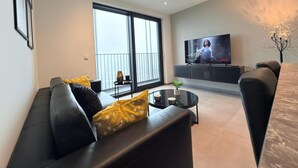Een moderne woonkamer met een zwarte leren bank, een glazen salontafel en een grote flatscreen-televisie op een zwart voetstuk.