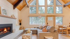 A cozy living room with a fireplace, large windows, a sofa, and a dining area. - Peaceful Tahoe Retreat by Avantstay Cozy Modern Cabin Sauna, Hot Tub + Fireplace Near Lake (Carnelian Bay)
