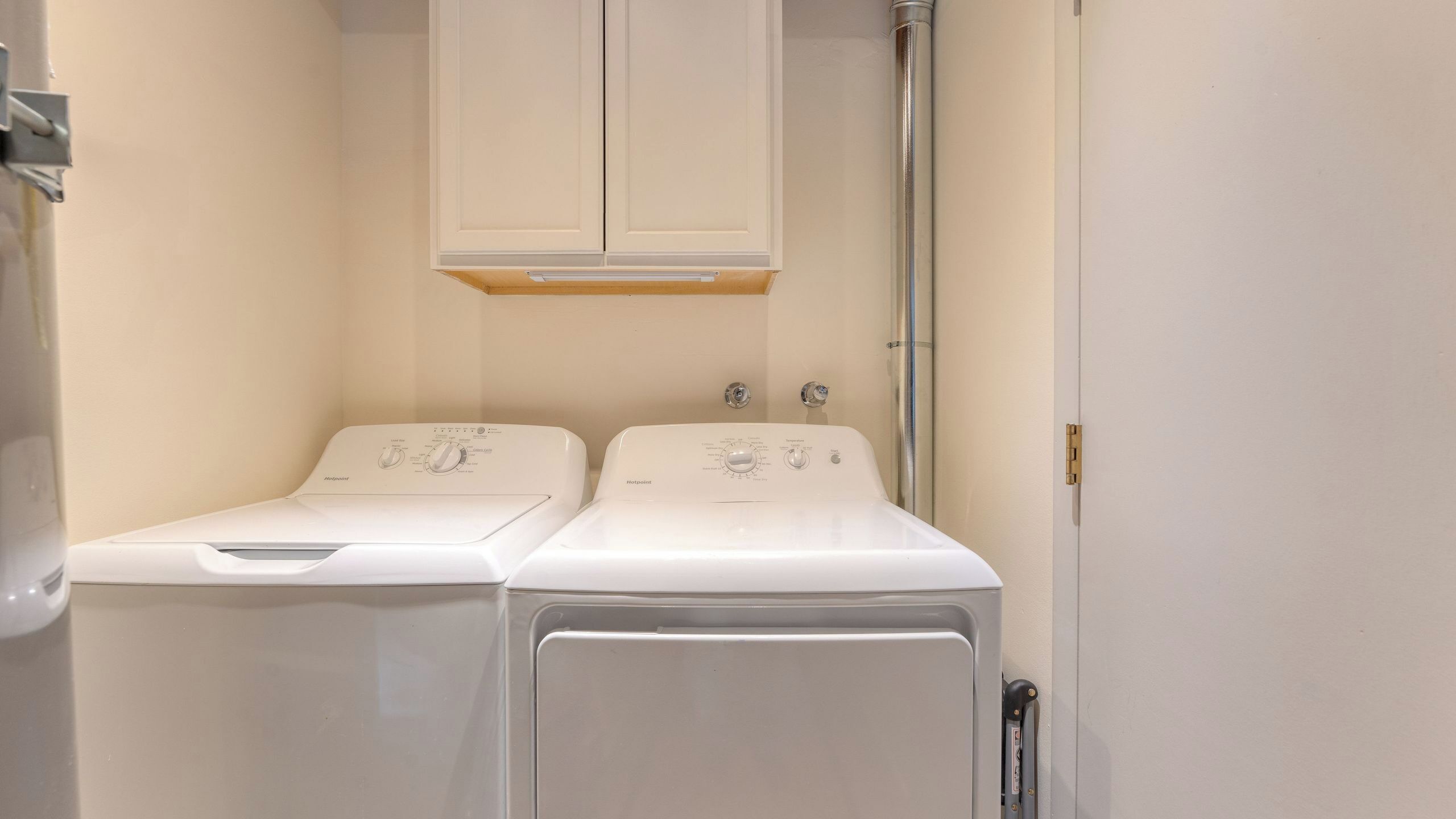 A laundry room with two white front-loading washers and dryers, a metal pipe, and a wooden shelf above the dryers.