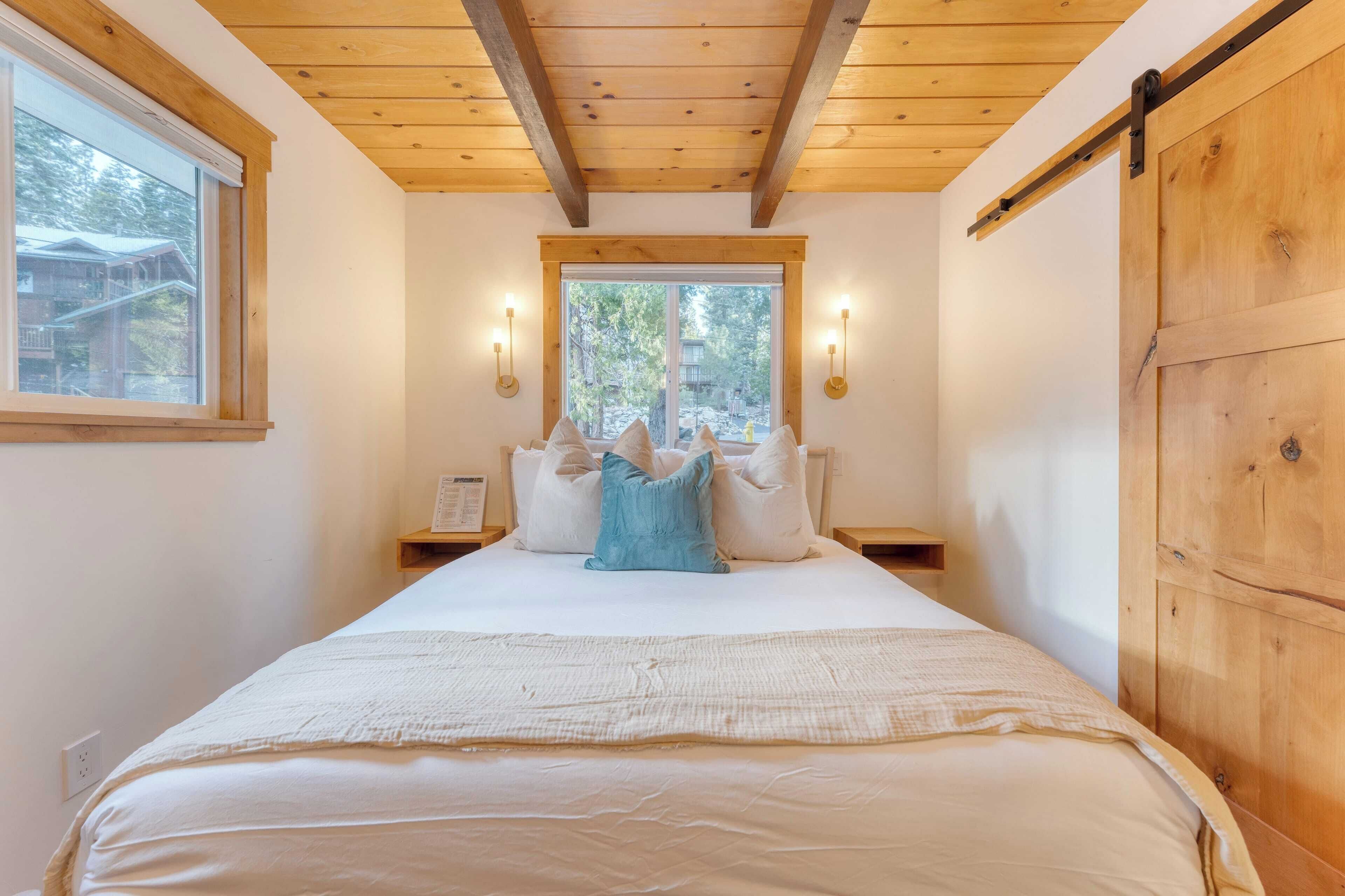 A bedroom with a large bed, wooden ceiling beams, a window with a view of trees, and two bedside lamps.