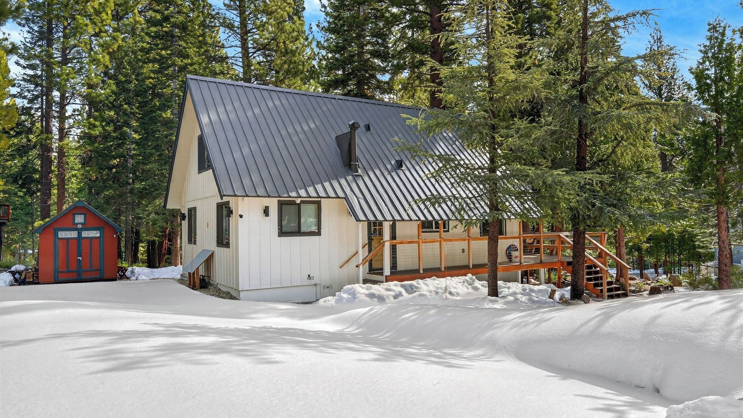 A cabin with a metal roof, wooden staircase, and a red shed in a snowy forest.