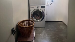 A laundry room with two stacked front-loading washing machines, a wicker chair, and a small window providing natural light. - Keepsake Oasis, Mid-Island 3-Bed Modern Cozy Home with AC, WiFi, Quiet Residence (Christiansted)