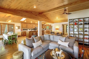 A spacious living area with a sectional sofa, a dining table with chairs, a kitchen in the background, and a bookshelf filled with various items. - Short Drive to Itasca State Park - Waterfront Home (Park Rapids)