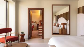 Habitación de hotel con una cama, una silla, una mesita con plantas, un espejo y vistas a una escalera que lleva a un baño.