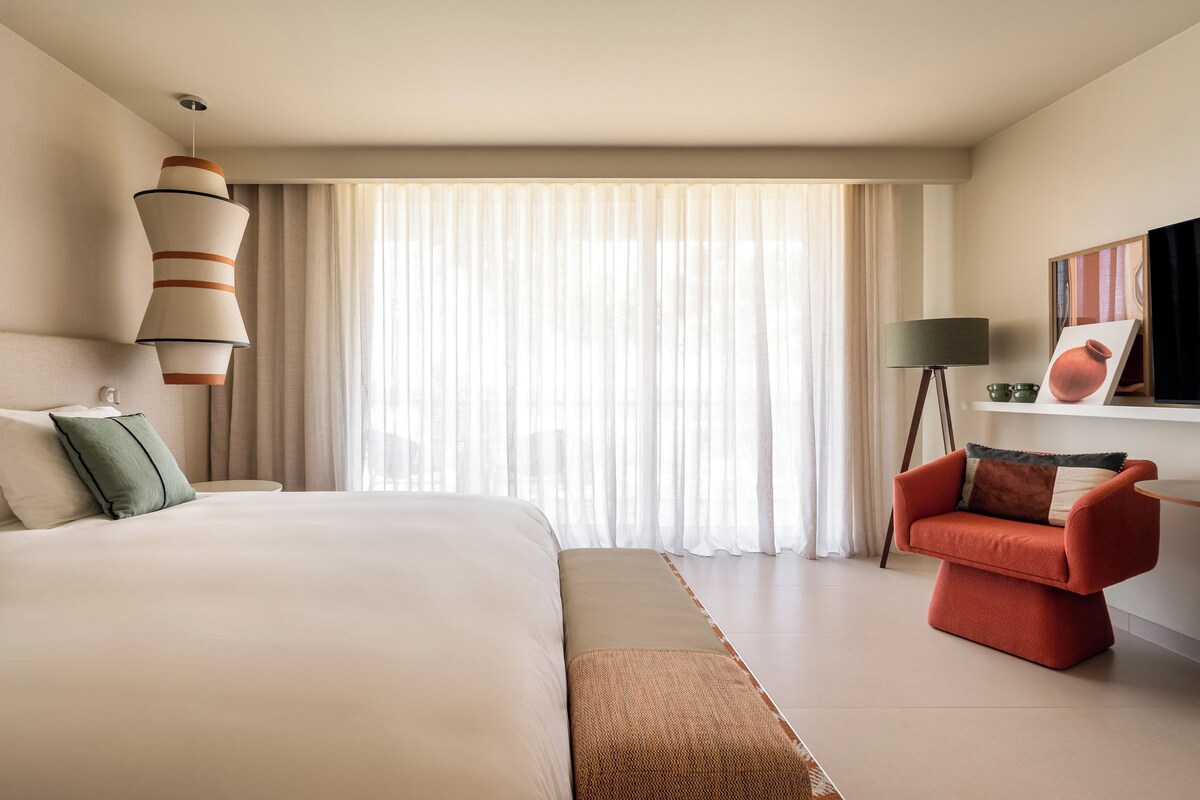 A modern hotel room with a large bed, a red armchair, a standing lamp, and a painting on the wall.