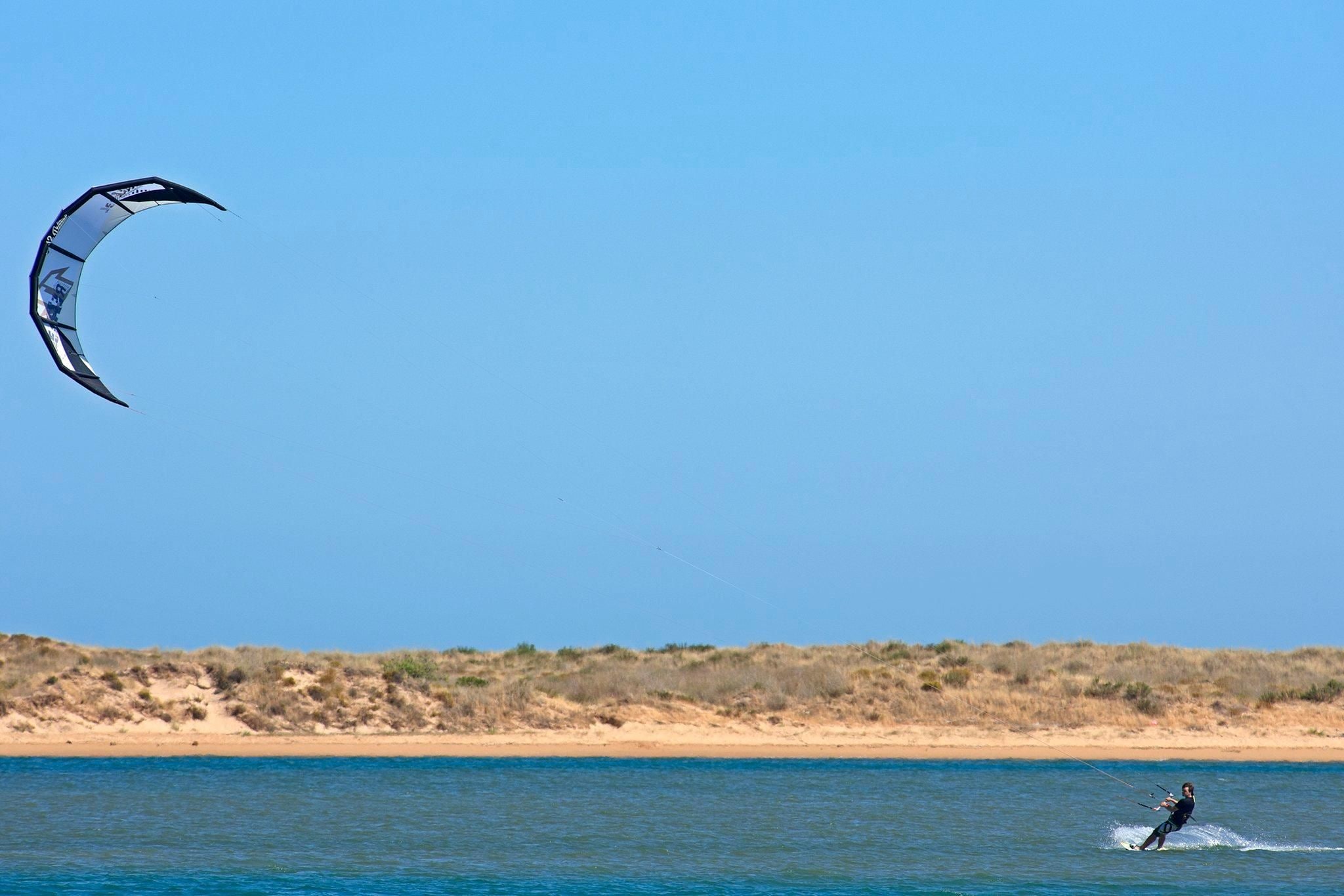 Una persona practica windsurf en una playa bajo un cielo despejado.