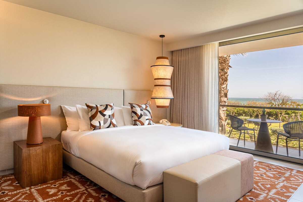 A hotel room with a large bed, a nightstand with a lamp, a view of the outdoors through a balcony, and a small table with chairs.