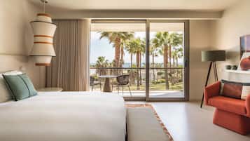 Quarto de hotel com cama, sofá, mesa e vista para palmeiras e o oceano.