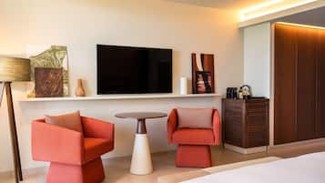A modern hotel room with a bed, two red armchairs, a small round table, a TV, and a wardrobe.
