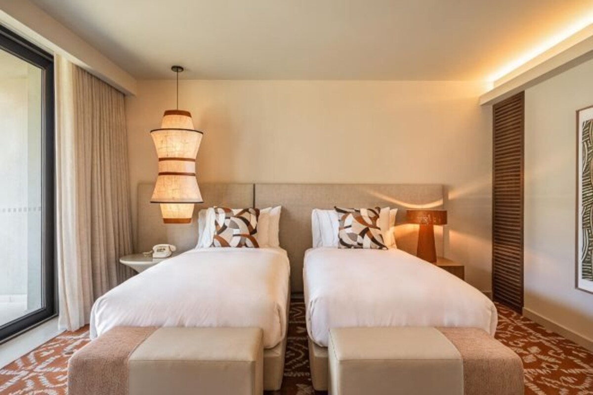 A hotel room with two beds, a bedside table with a lamp, a phone, and a large window with curtains.