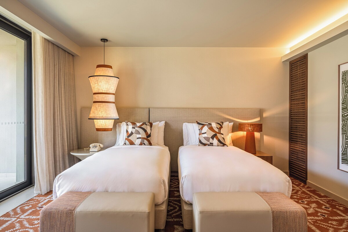 A hotel room with two beds, a bedside table with a lamp, a telephone, a window with curtains, and a patterned carpet.