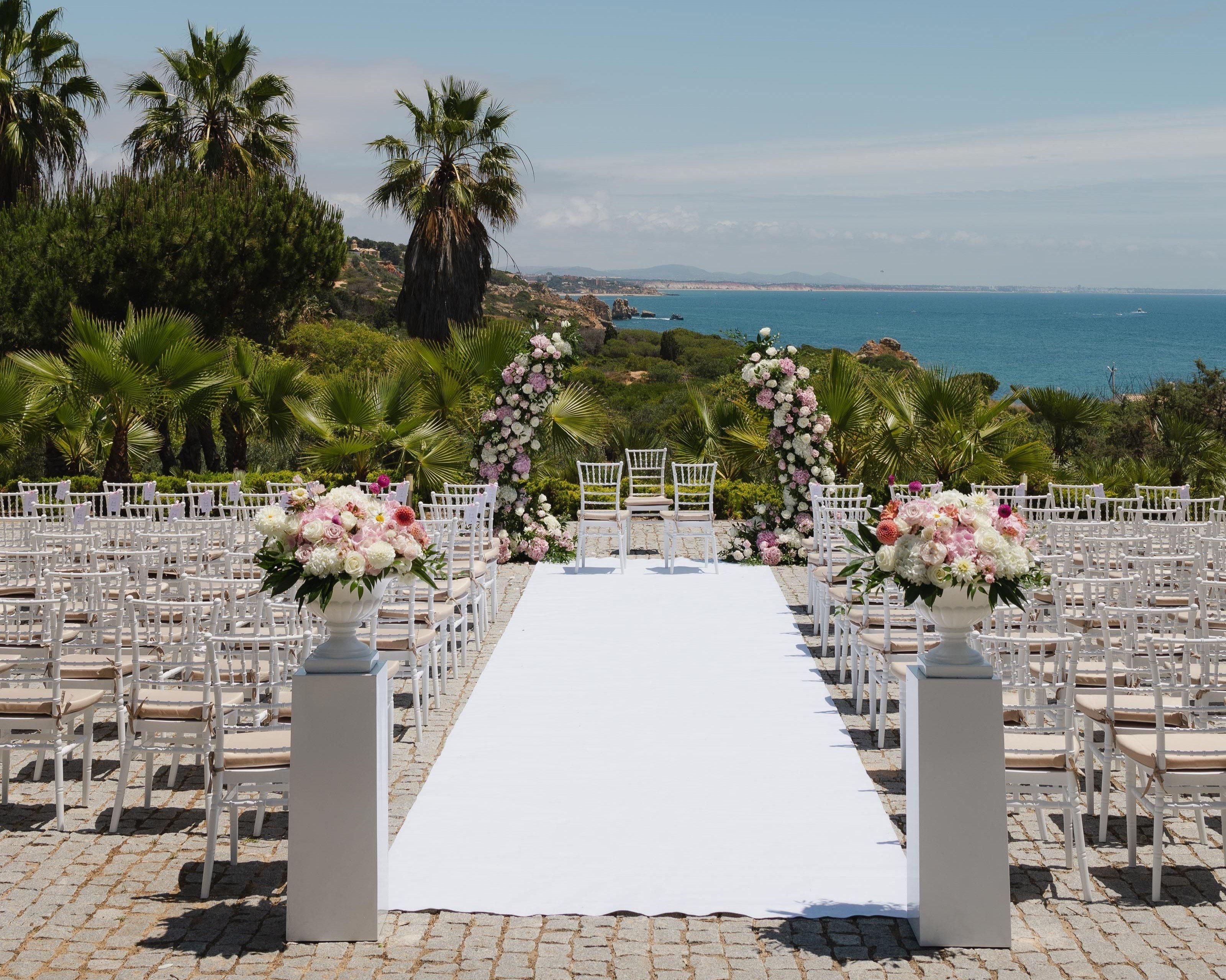 Un pasillo nupcial con sillas blancas y una alfombra blanca, que conduce a un arco decorado con flores.