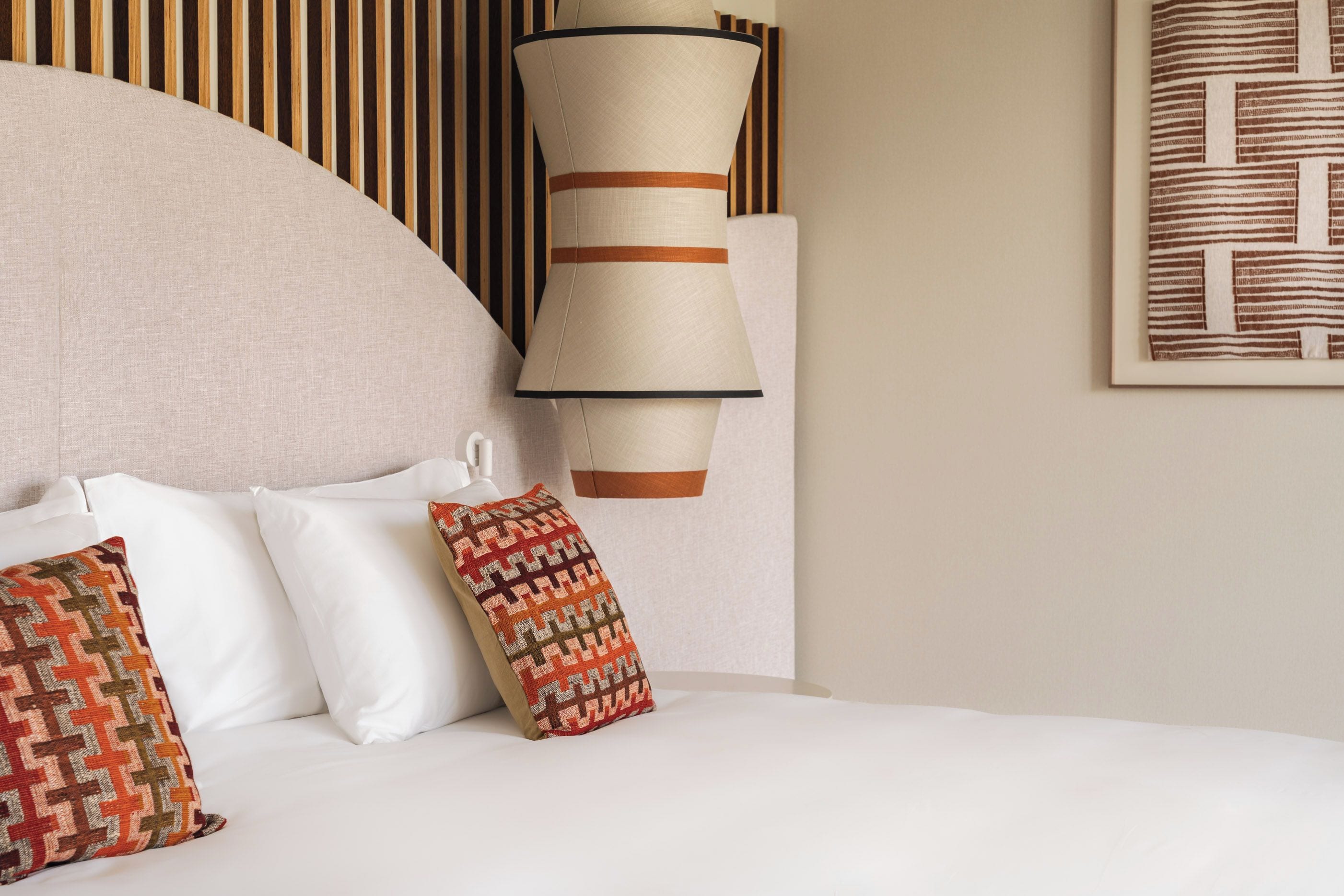 Una cama con ropa blanca, un cojín estampado y una lámpara de pared.