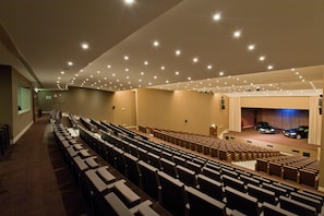 An auditorium with rows of seats, a stage with a blue curtain, and ceiling lights. - Kimpton Atlantico Algarve (Albufeira)
