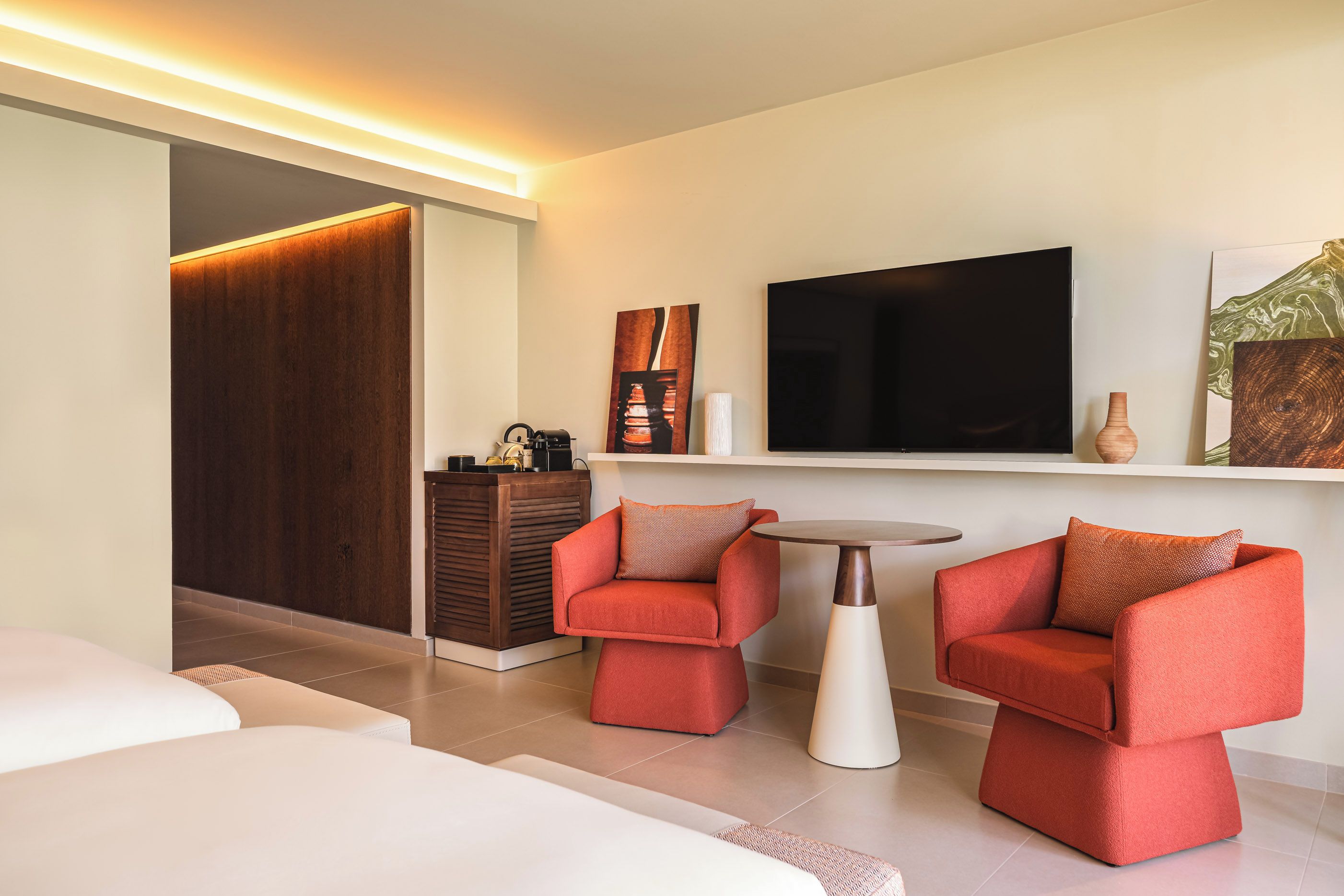 Een moderne hotelkamer met een televisie, twee rode fauteuils, een klein rond tafeltje en een houten garderobe.