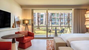 Habitación de hotel con cama, sofá rojo, televisión, lámpara y vista a un patio con palmeras y un edificio.