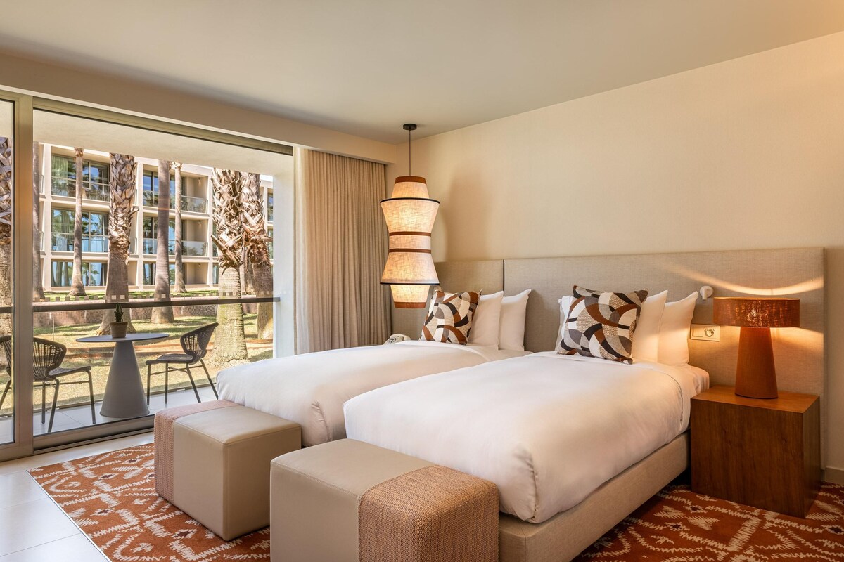 A hotel room with two beds, a balcony with a dining area, and a view of a building.