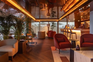 A modern interior with wooden flooring, red armchairs, a bar area, and large windows. - Kimpton Atlantico Algarve (Albufeira)