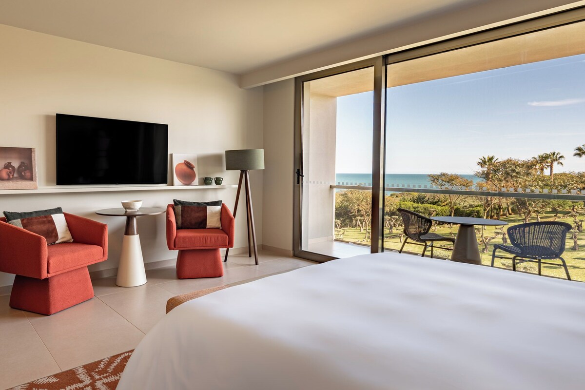 A hotel room with a bed, two red sofas, a TV, a lamp, a small table, and a balcony with chairs and a view of the sea.