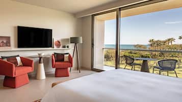 Une chambre d’hôtel avec un lit, deux canapés rouges, une télévision, une lampe, une petite table et un balcon avec des chaises et vue sur la mer.