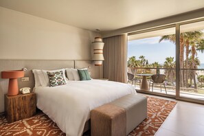 Une chambre d’hôtel moderne dotée d’un grand lit, d’un téléphone sur la table de chevet, d’un tapis à motifs et d’un balcon offrant une vue sur les palmiers et l’océan.