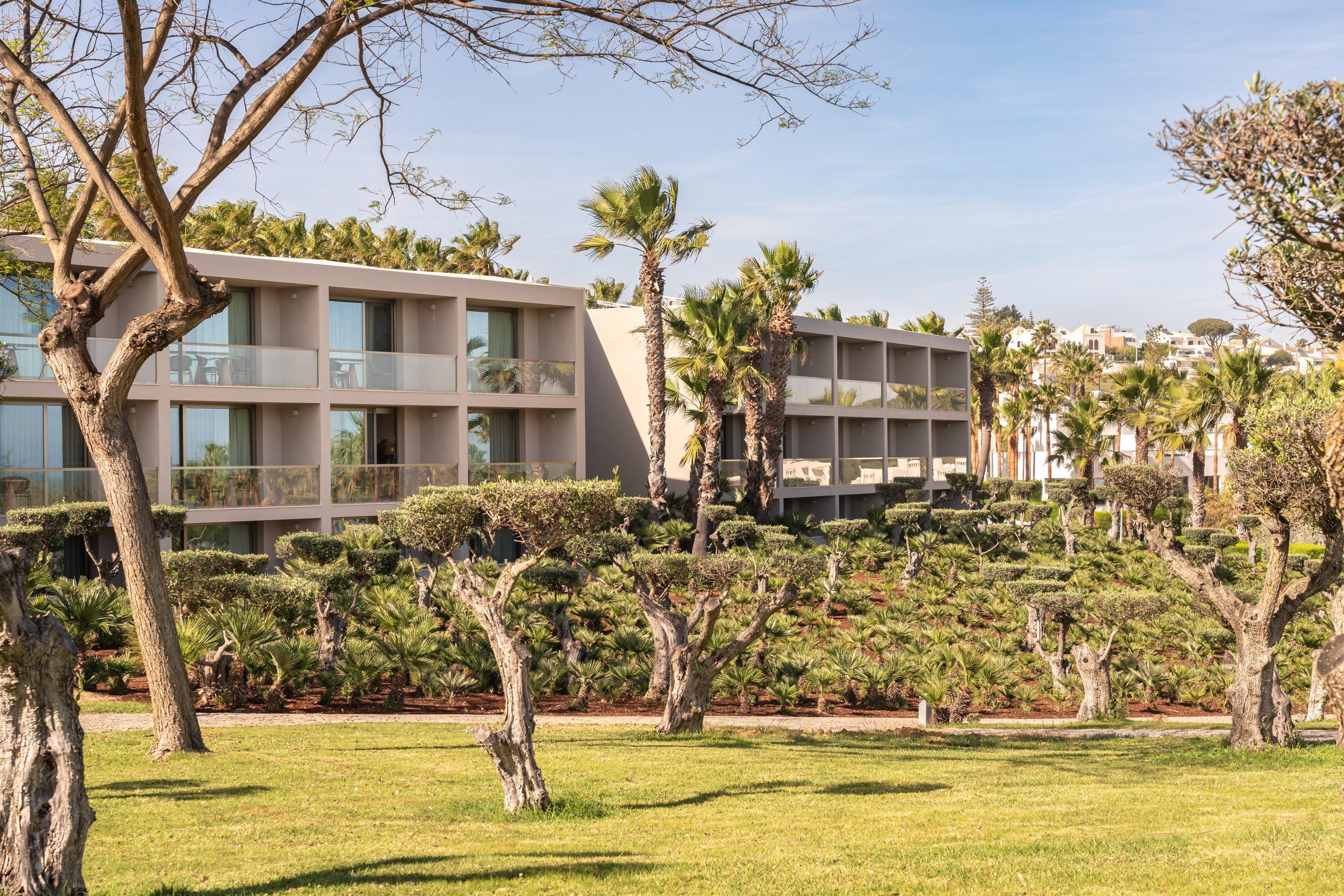 Un hôtel moderne à plusieurs étages, doté d’un jardin bien entretenu avec différentes espèces d’arbres, et offrant une vue dégagée sur les environs.