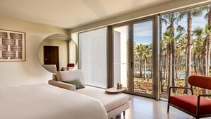 A hotel room with a bed, a sofa, a chair, a small table, and a large window with a view of palm trees. - Kimpton Atlantico Algarve (Albufeira)