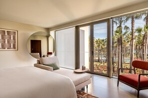 A hotel room with a bed, a sofa, a chair, a small table, and a large window with a view of palm trees. - Kimpton Atlantico Algarve (Albufeira)