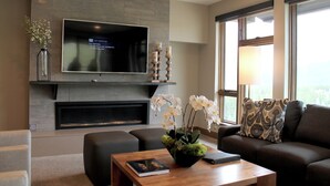 Una sala de estar moderna con un televisor de pantalla plana, una chimenea, una mesa de centro con un libro y un jarrón de flores, y un sofá con cojines estampados.