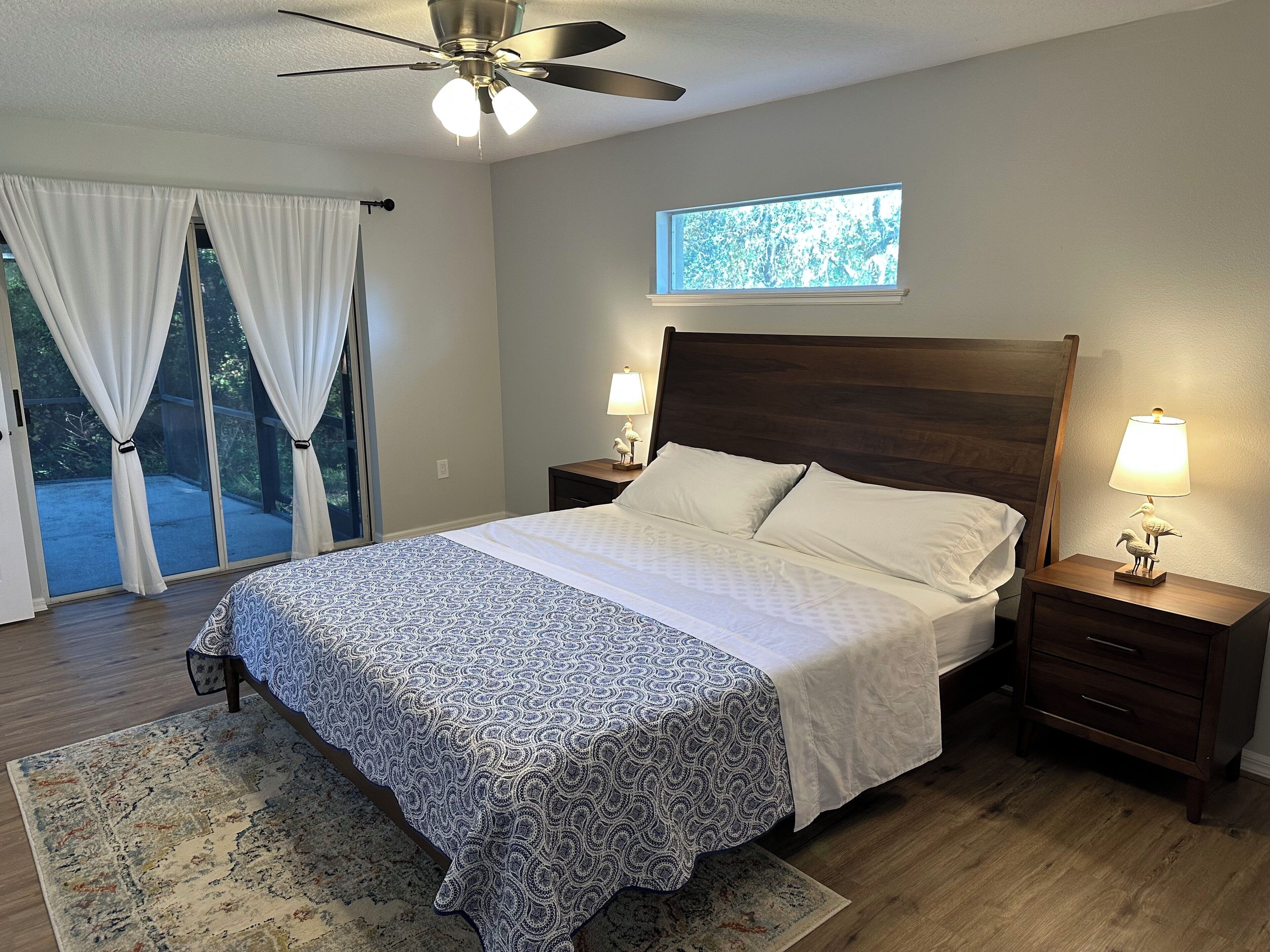 A bedroom with a bed, two bedside tables with lamps, a ceiling fan, a window with a view, and a sliding glass door leading to an outdoor area.