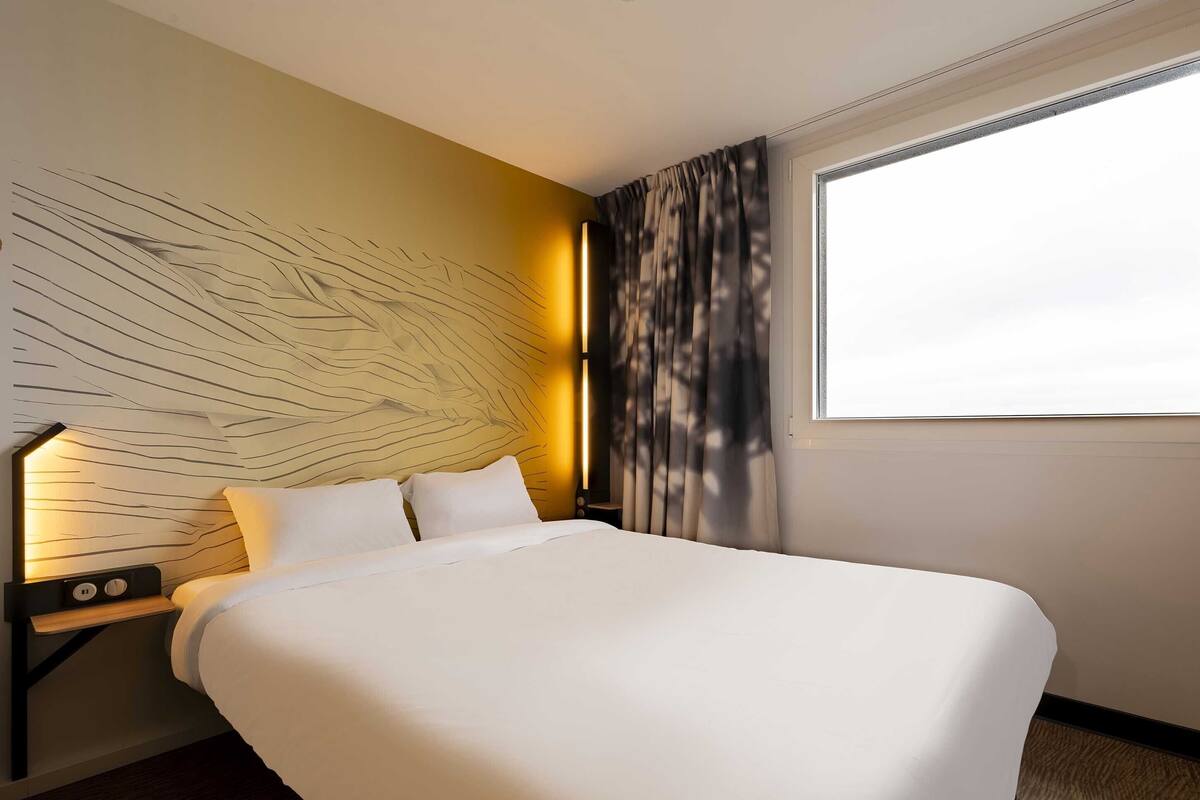 Hotelkamer met een groot bed, nachtkastje met lamp, wand met een uniek ontwerp en raam met gordijnen.