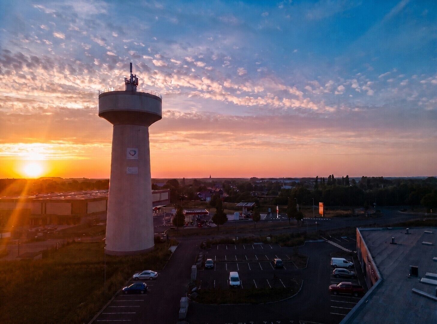 Una torre cilíndrica grande con la parte superior plana, rodeada por un estacionamiento con varios autos, con el cielo al atardecer como fondo.