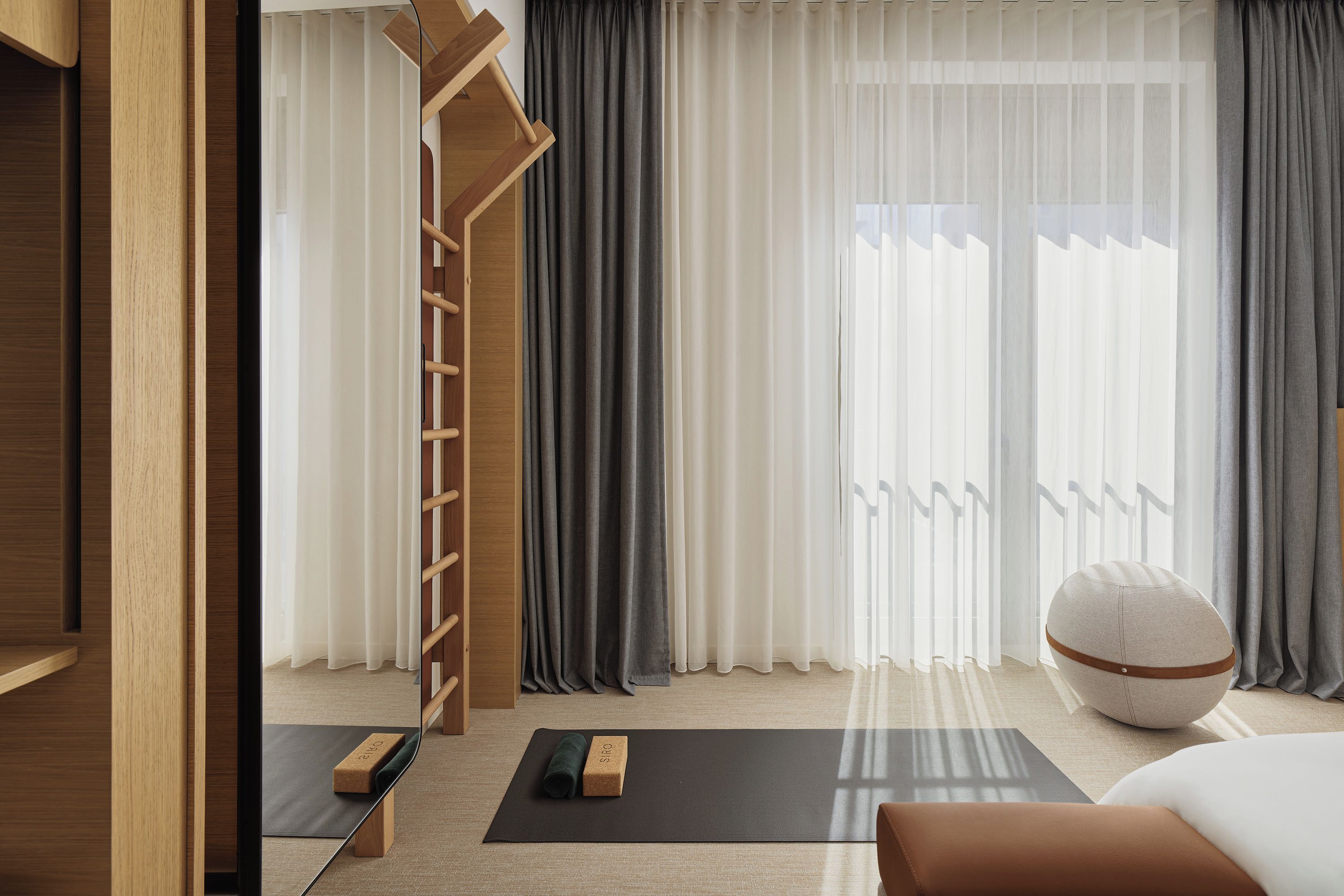 A modern hotel room with a wooden floor, a yoga mat, a small wooden stool, a round ottoman, and a bed.