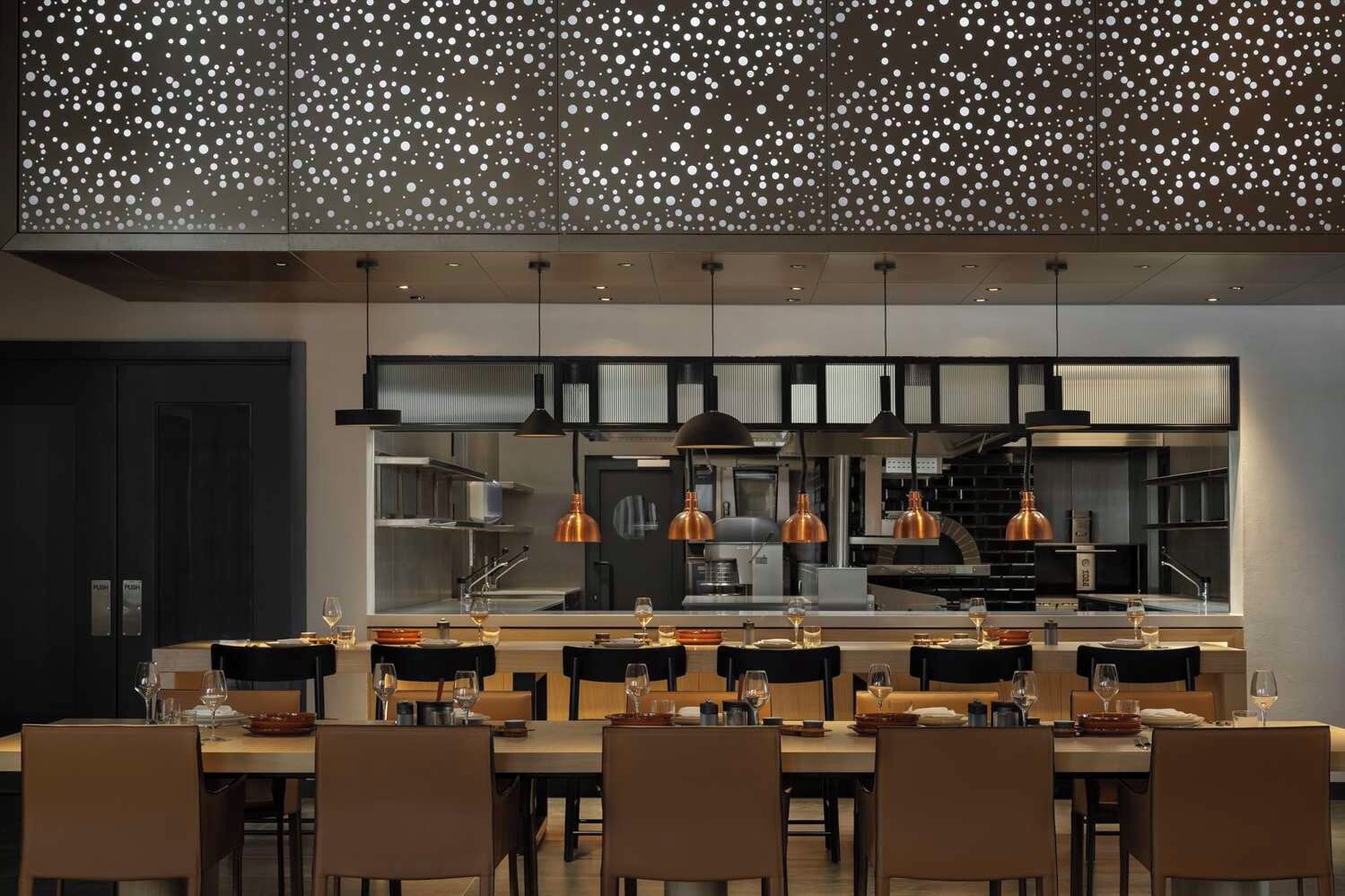 Un restaurante con una decoración moderna, que presenta una larga barra con varias mesas y sillas, iluminación colgante y una cocina a la vista.