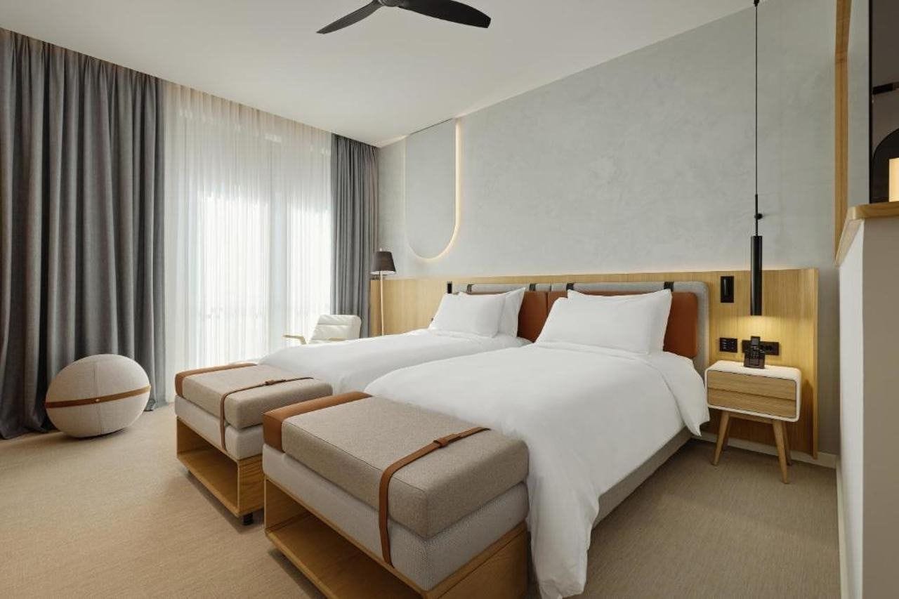 A hotel room with two beds, a bedside table, a lamp, a chair, and a large window with curtains.